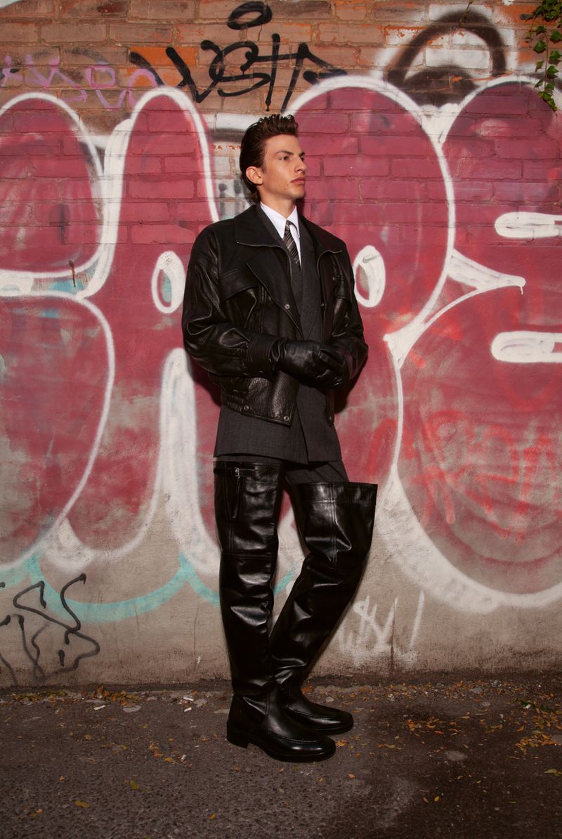 A young man stands tall in a leather jacket, suit, and thigh-high boots against a graffiti-covered brick wall in an outdoor urban setting.