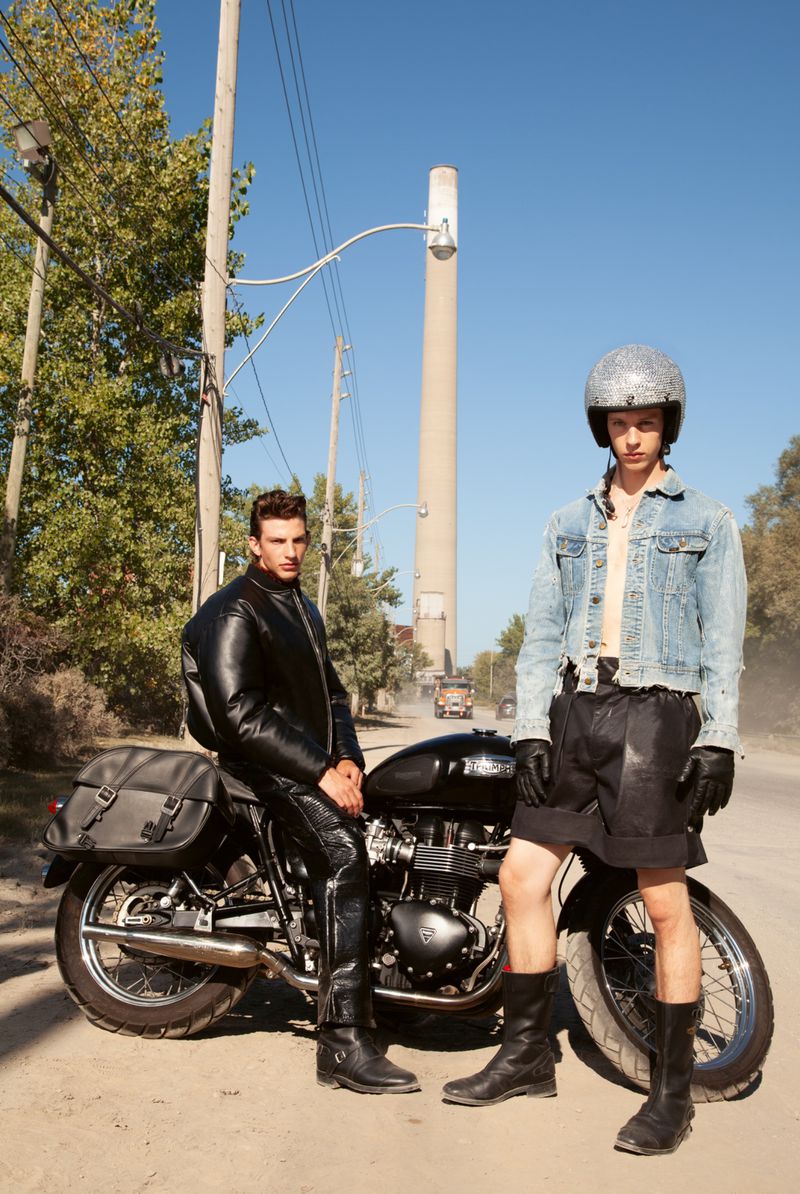 Two men stand with a black motorcycle on a dirt road surrounded by trees under a clear sky, one wearing a motorcycle helmet.