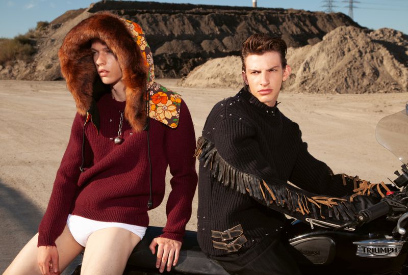 Two adult male fashion models pose next to a motorcycle on a dirt road with hills in the background, wearing fashionable knitwear and accessories.