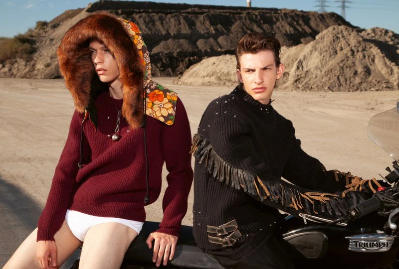 Two adult male fashion models pose next to a motorcycle on a dirt road with hills in the background, wearing fashionable knitwear and accessories.