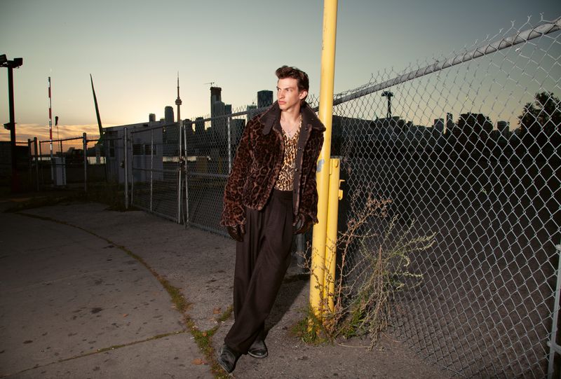 A young man in a leopard print jacket and shirt poses against a chainlink fence in an urban setting during twilight.