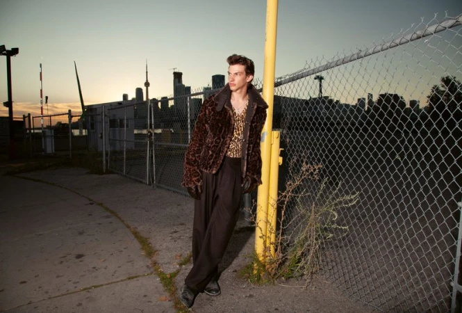 A young man in a leopard print jacket and shirt poses against a chainlink fence in an urban setting during twilight.