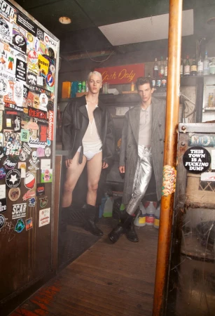 Two male models posing inside a dimly lit bar covered in stickers, wearing coats and casual wear.