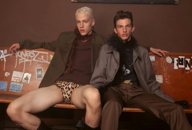 Two young male models seated together on a graffiti-covered wooden bench for an editorial feature.
