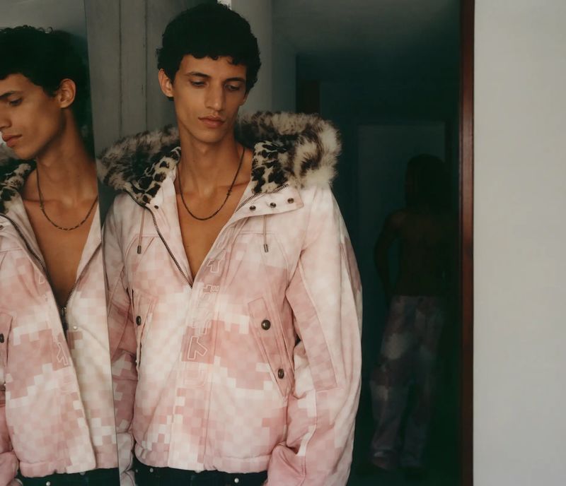 Young man in a pink camouflage hooded jacket standing indoors, reflected in a mirror, fashion portrait, studio lighting.