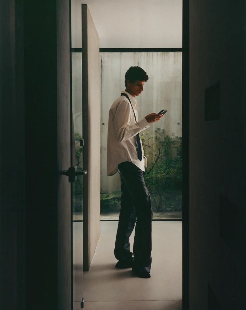 A young adult stands indoors, using a smartphone in a white shirt and black pants, near a doorway with a glimpse of greenery outside.
