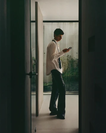 A young adult stands indoors, using a smartphone in a white shirt and black pants, near a doorway with a glimpse of greenery outside.