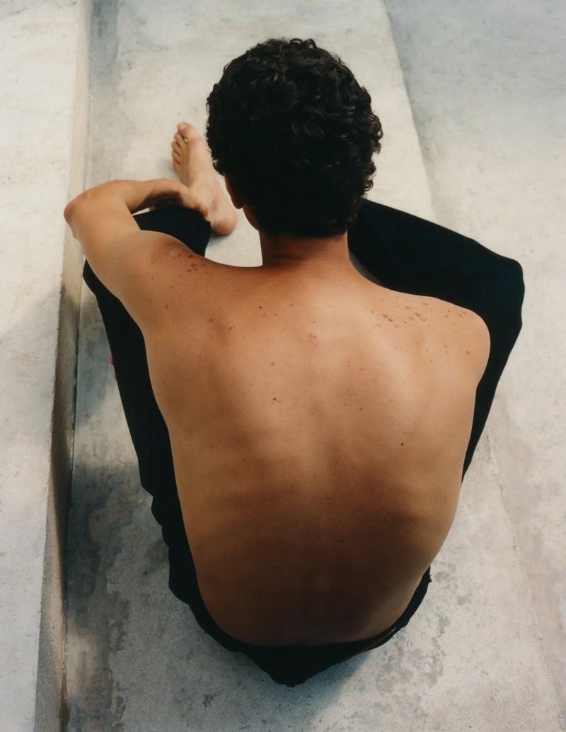 High angle view of a shirtless man with dark curly hair sitting on concrete steps with his back to the camera, showcasing his back and shoulders.