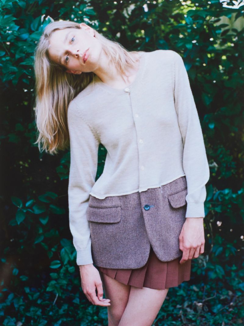 A fair-skinned woman with blonde hair is outdoors modeling a neutral-colored cardigan and tweed mini skirt against a lush green leafy background.