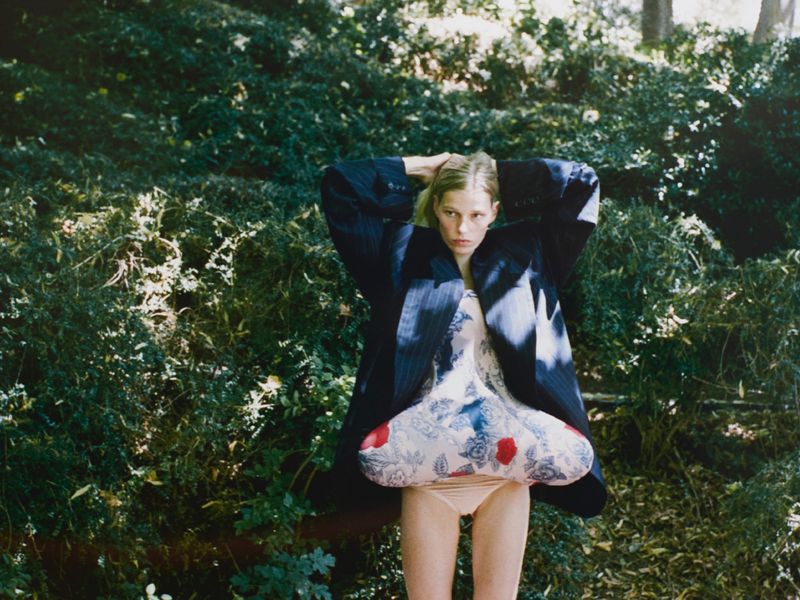 Fashion shot of a fair-skinned adult female model in a dress with a blazer, posing in a lush outdoor wooded environment with filtered sunlight.