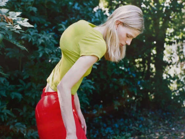 Model wearing a chartreuse short-sleeve top and red skirt leaning forward outdoors amid green foliage