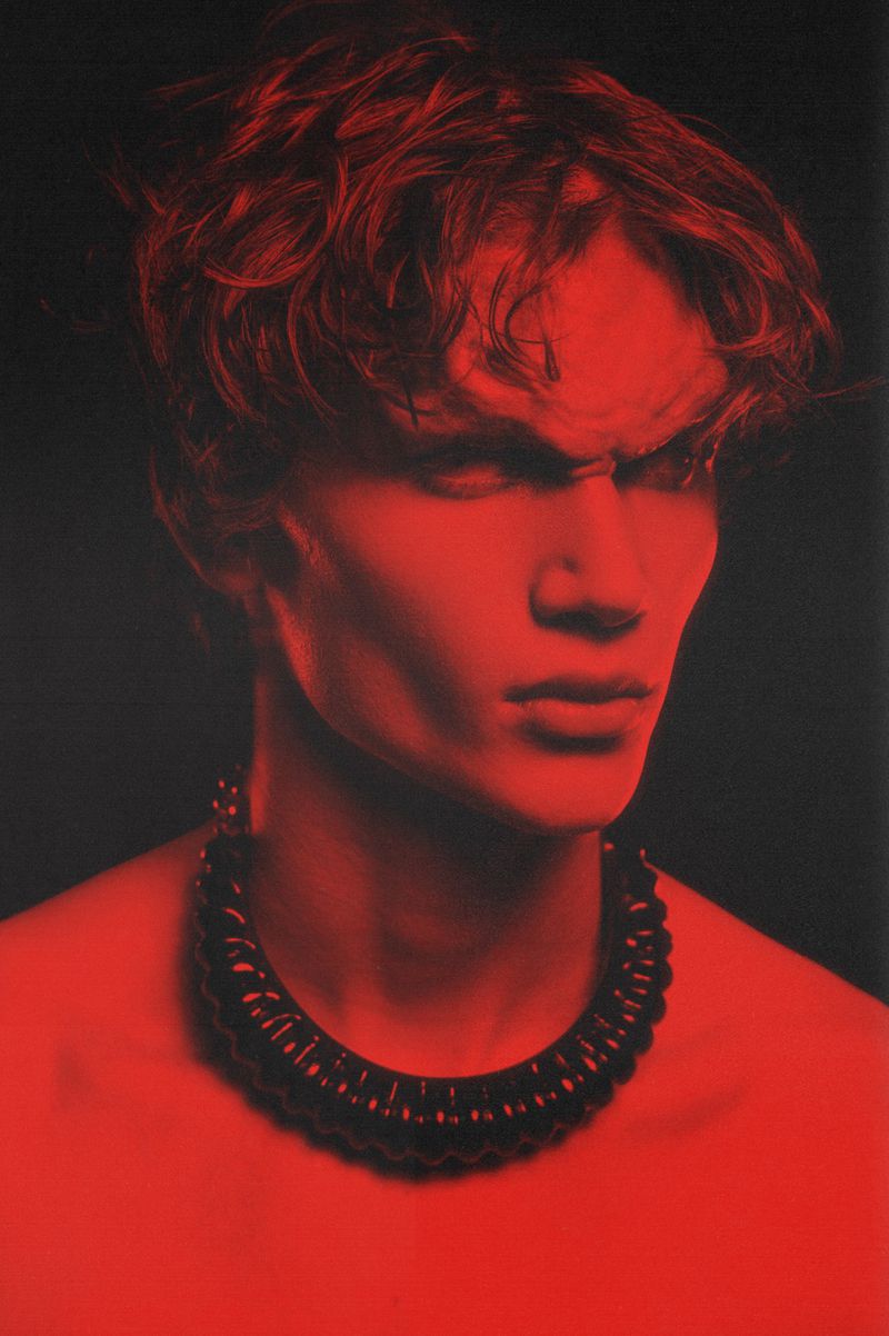 Close-up portrait of a young adult male model with red lighting and a sculptural necklace against a dark background, fashion editorial.
