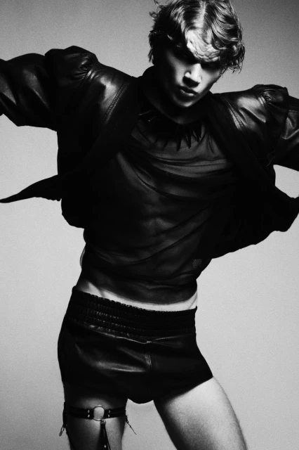 Monochrome portrait of a young adult male fashion model wearing leather shorts and black top, posed in a photo studio setting.