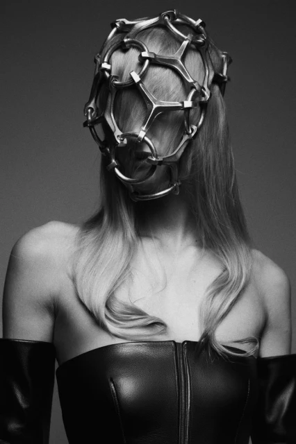 Monochrome studio shot of a woman wearing a sculptural metal headpiece, black corset top, and matching arm coverings against a grey backdrop.