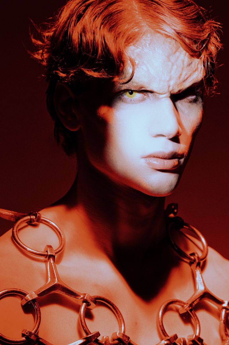 Close-up portrait of a person with dramatic red hair, light skin and reptile-like facial features, wearing a metal neck piece.