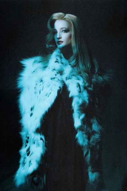 A woman with long blond hair poses in front of a dark background while wearing a fur stole. The image is lit with a cool, blue tone.