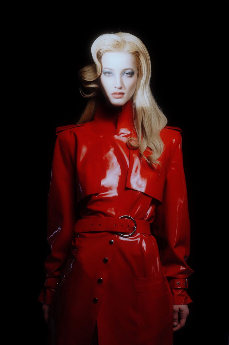 A woman with blonde hair poses in a shiny red vinyl trench coat against a dark background in a fashion or beauty style photo.