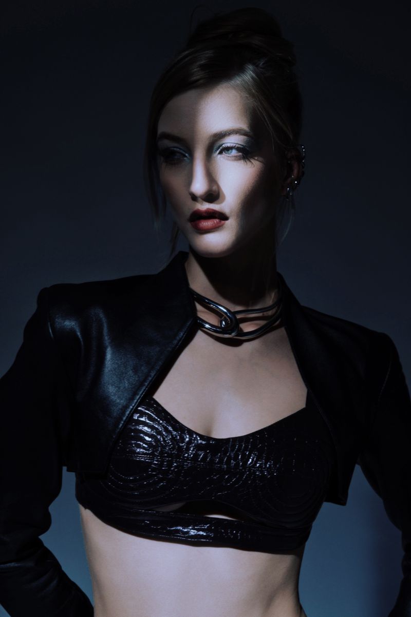 Fashion shot of a woman with dark hair in a high bun, wearing a leather jacket and a metal necklace in a dark photo studio.