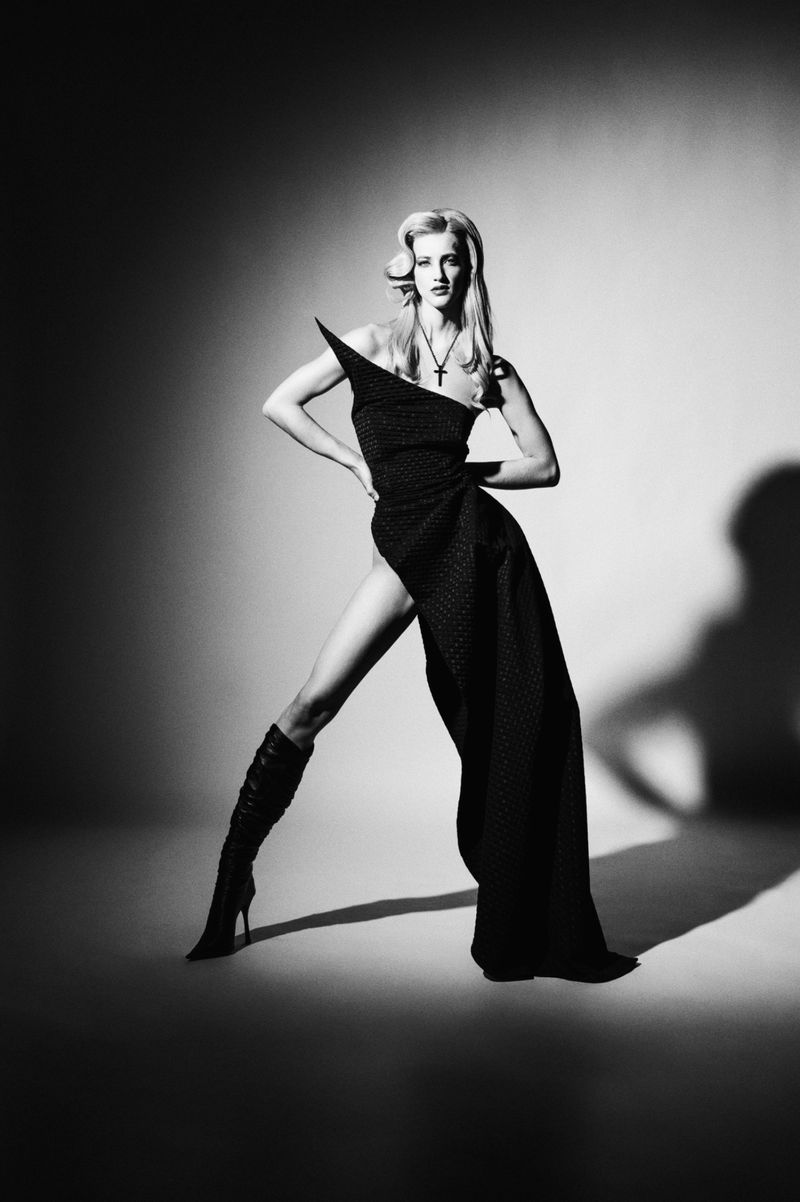 Fashionable woman posing in a black dress and boots against a grey background with dramatic lighting in a studio setting.