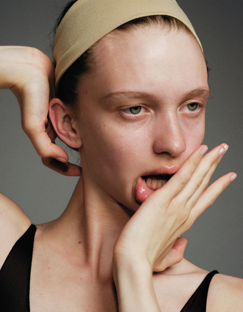 Close-up studio portrait of a young adult woman with a neutral expression, wearing a beige headband, and posing with her hand on her face.