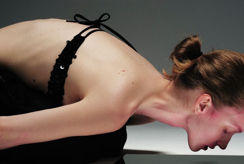 Bare shouldered woman in a black beaded bra top kneeling, looking down with a neutral expression in a neutral gray photo studio setting.