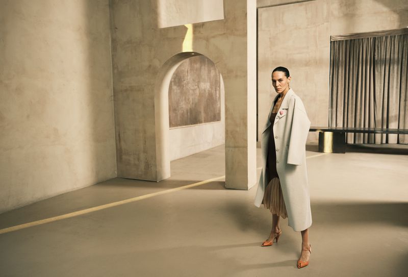 Fashionable adult woman stands in modern space wearing a long coat and dress with heeled shoes, posing near structural elements and drapery.