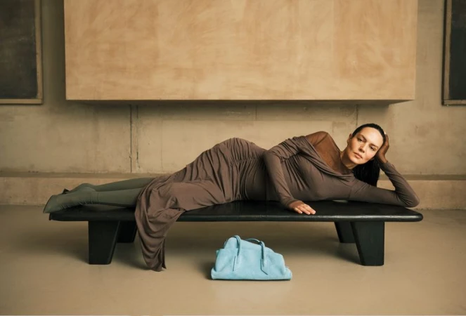 Model reclining on a black wooden bench wearing a ruched brown dress with a light blue handbag placed underneath.