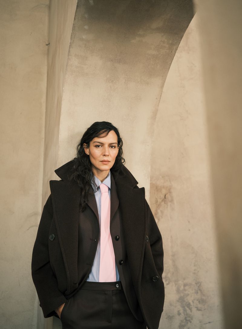 Woman in brown coat and suit poses indoors in front of archway; light blue shirt with a pink tie, and curly dark hair.