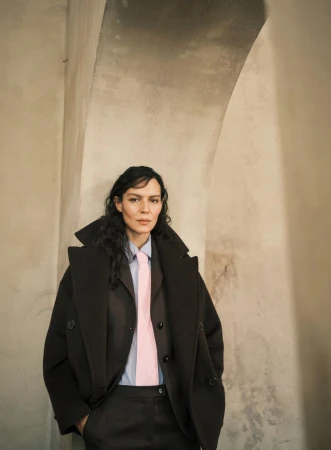 Woman in brown coat and suit poses indoors in front of archway; light blue shirt with a pink tie, and curly dark hair.