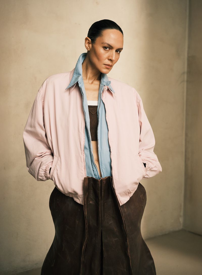 Adult female wearing a pink jacket, brown crop top, and brown skirt in a studio with a neutral color palette in a portrait shot.