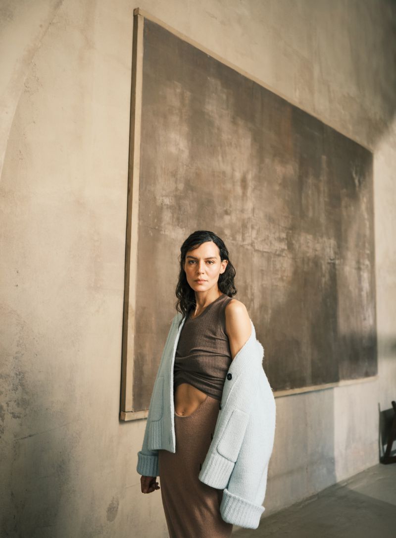 A woman in a sleeveless top and skirt with a light-blue jacket is standing indoors in front of a framed abstract artwork.