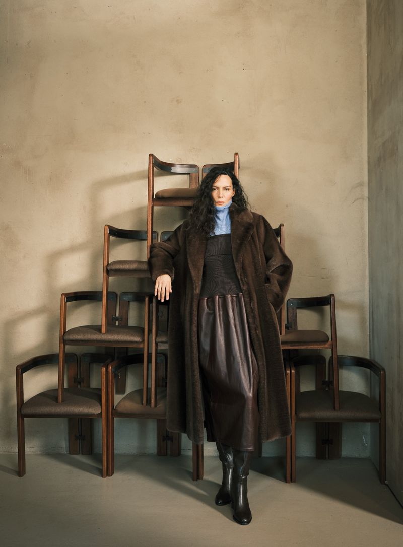 A brunette woman in a brown coat poses next to a stack of wooden chairs in a studio setting, creating a fashion-forward editorial image.