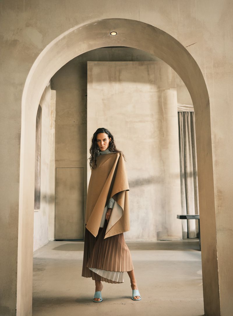 A woman posing, wearing a turtleneck, a tan poncho, a pleated skirt, and high-heeled sandals stands in an arched hallway.