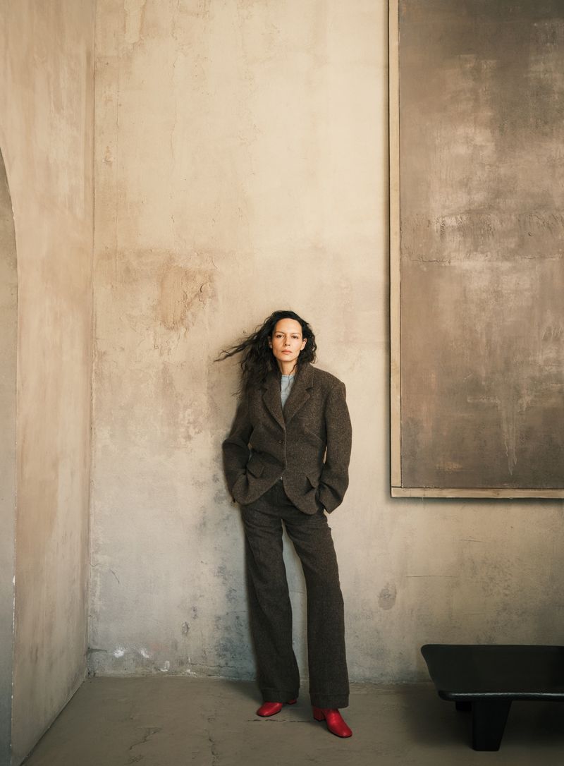 A woman in a brown suit and red shoes leans against a textured wall with modern art in a naturally lit indoor setting.