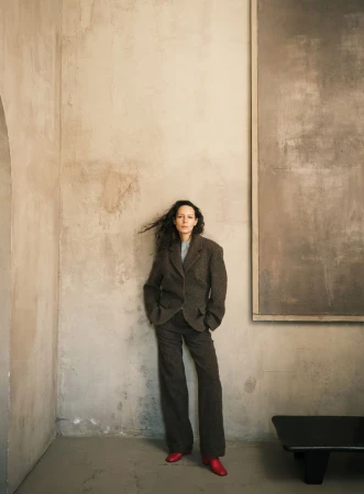 A woman in a brown suit and red shoes leans against a textured wall with modern art in a naturally lit indoor setting.