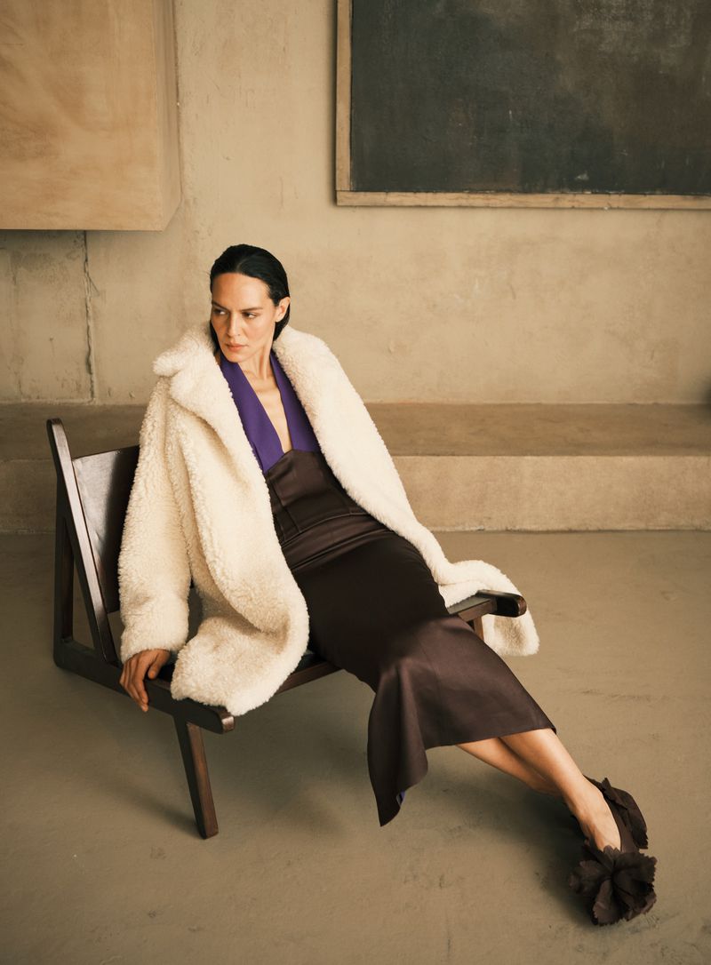 A woman in a brown dress and cream-colored coat is sitting on a wooden chair in a neutral-toned room with art hanging on the wall.