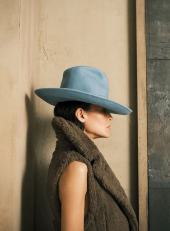 Profile shot of an adult woman wearing a wide-brimmed baby-blue hat and a brown, furry vest against a textured beige wall and blue panel.
