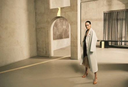 Model wearing a long beige coat and orange heels in a minimal concrete architectural interior for Vanity Fair Italia.