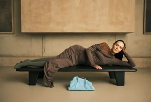 Model reclining on a black wooden bench wearing a ruched brown dress with a light blue handbag placed underneath.