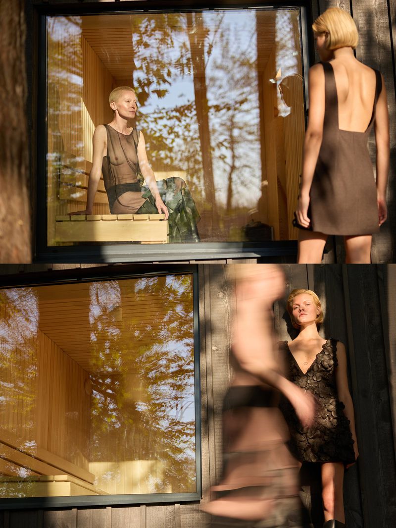 Two women pose near a sauna, one inside a glass window and another beside it, with the background showing a forest. 