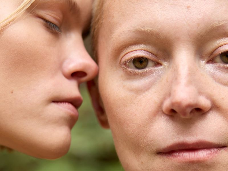 Close-up portrait of two fair-skinned women, one with closed eyes and the other looking directly at the camera against a green background.