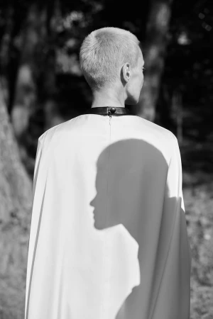 Woman with a shaved head wearing a light colored cape and a black collar standing in a dark forest in black and white.