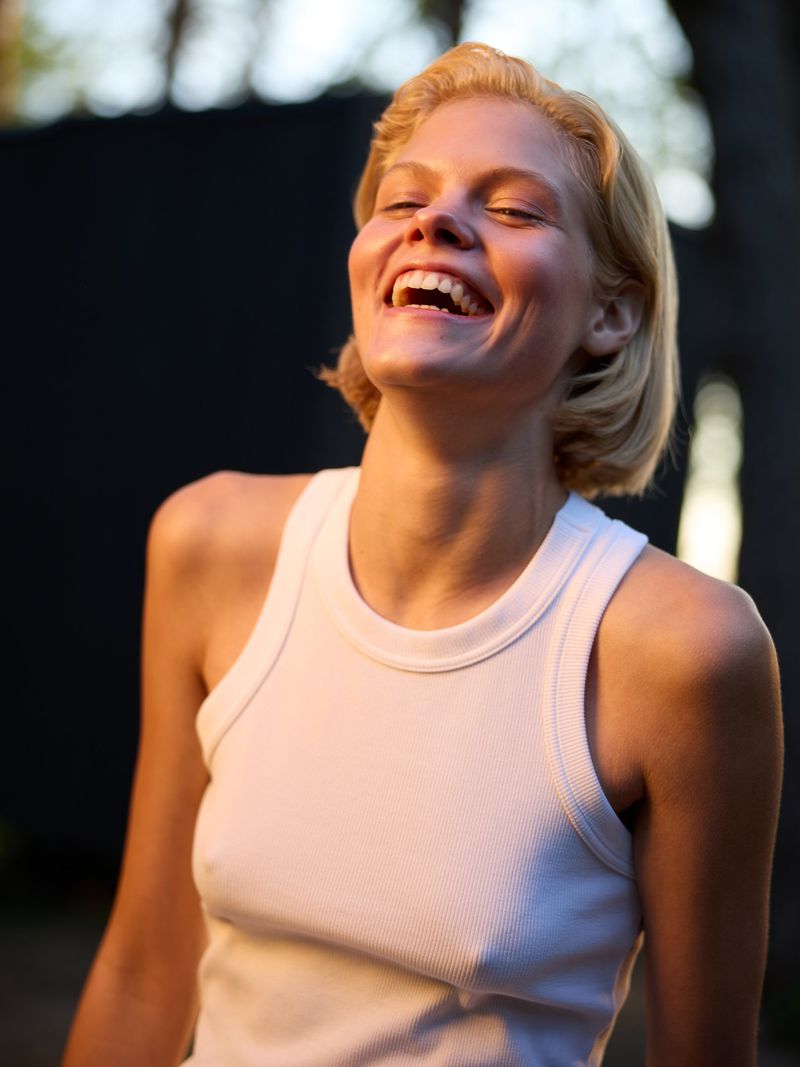 Close-up of a smiling young adult woman with short blonde hair wearing a white tank top, set against a blurred outdoor background.