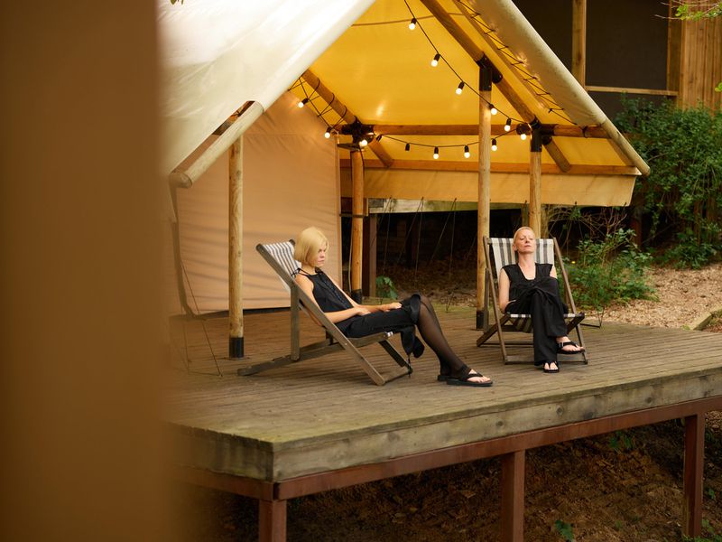 Two women in black outfits sit in deck chairs on a wooden platform outside a glamping tent with string lights, surrounded by lush greenery.