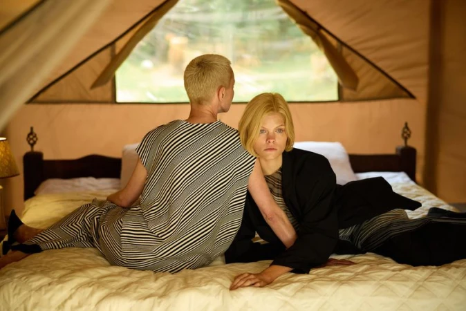 Two fair-skinned adults sit on a bed inside a tent. One wears a striped top, while the other wears a black jacket and patterned shirt.