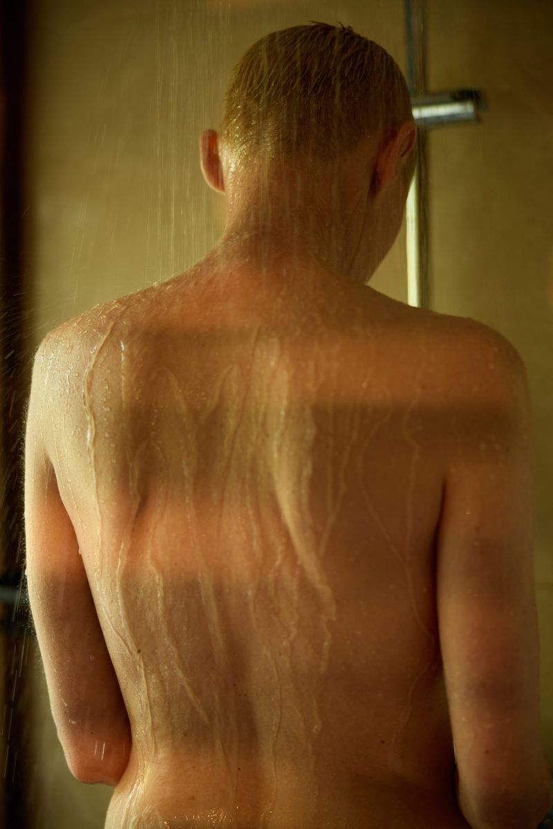Back view of an adult taking a shower with water streaming down in a warmly lit bathroom with beige tile and chrome shower head.