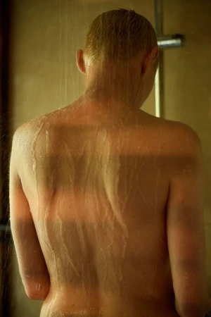 Back view of an adult taking a shower with water streaming down in a warmly lit bathroom with beige tile and chrome shower head.