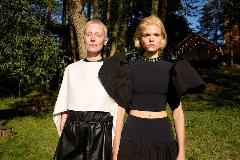 Two adult women in fashionable black and white outfits pose outdoors on a sunny day, with a blurred forest and cabin in the background.
