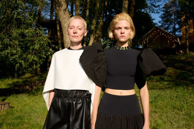 Two adult women in fashionable black and white outfits pose outdoors on a sunny day, with a blurred forest and cabin in the background.