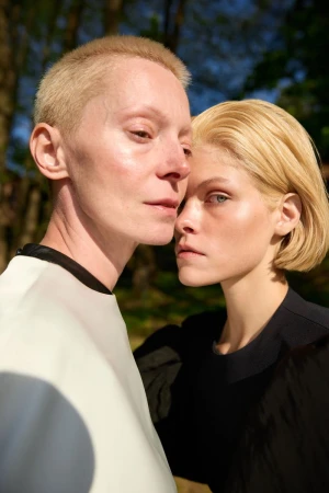 Two adult women with blonde hair pose closely together outdoors in black and white clothing in a portrait with a blurred background.